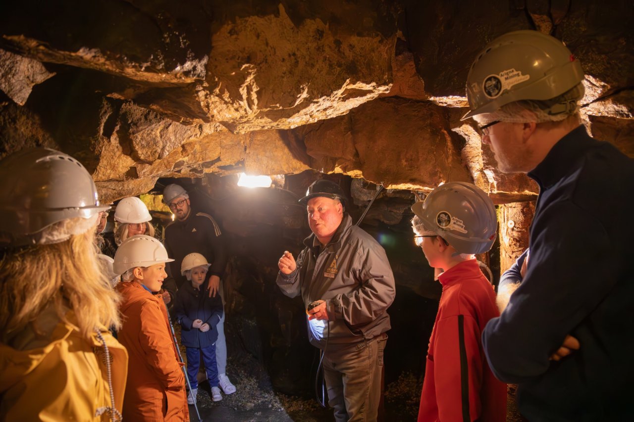 Guests gathered around guide listening as he tells the history of the Arigna Mines. Each person is wearing warm clothes and a safety helmet. 